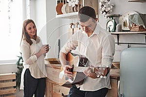 Girl is smiling. Young guitarist playing love song for his girlfriend in the kitchen