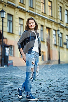 A girl in sneakers, jeans and a leather jacket stands on a city