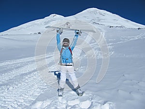 The girl-snowboarder stands in front of the mountain tops holding a snowboard over her head on a clear Sunny day