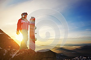 Girl snowboarder stands with snowboard on mountain`s top on sunset backdrop