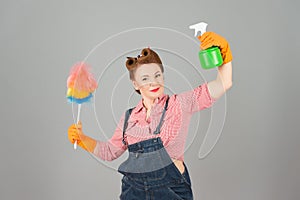 Girl with soft colorful duster and green spray bottle on grey background. Denim girl in orange gloves with brunette curls