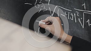 The girl solves the equations from the textbook at the blackboard. Close-up of a girl's hand writing on a blackboard