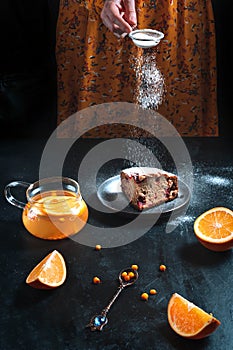 A girl sprinkles icing sugar on a piece of cake that stands on a dark table with tea.