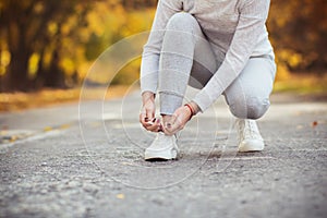 Girl squatted down to tie shoelaces on white sneakers on asphalt road, autumn sport concept outdoors