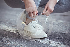 Girl squatted down to tie shoelaces on white sneakers on asphalt road, autumn sport concept outdoors