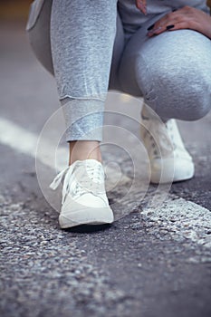 Girl squatted down to tie shoelaces on white sneakers on asphalt road, autumn sport concept outdoors