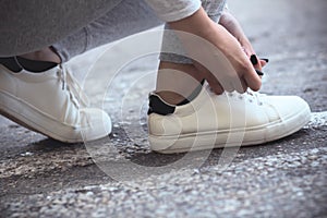 Girl squatted down to tie shoelaces on white sneakers on asphalt road, autumn sport concept outdoors