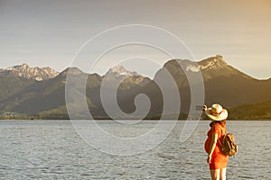 Girl with stalk hat and red dress on taking a selfie by a lake on a dock in front of big mountains
