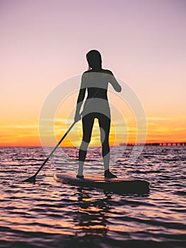 Girl stand up paddle boarding at dusk on a flat warm quiet sea with sunset colors