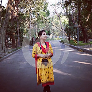 The girl is standing in the middle of a park street,she has a camera hanging around her neck.