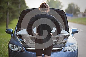 The girl is standing near the open hood of the car. The car needs repair. With smoke