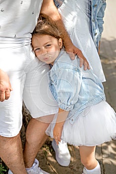 The girl is standing next to her parents with cute pouting lips and gently clinging to her father.