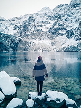 Girl is standing on the shore of a winter lake and mountains covered with snow