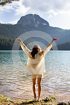 The girl stands with her back near a mountain lake in the background. Positive young woman traveling on a blue lake outdoors,