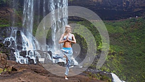 Girl stands on left leg folds palms in prayer by waterfall