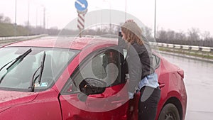 Girl stands near the broken car in the rain, she is wounded and holds her head.