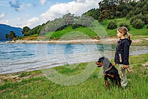 Girl stands near dog of Rottweiler breed in meadow next to lake, against hilly valley with spruce forests