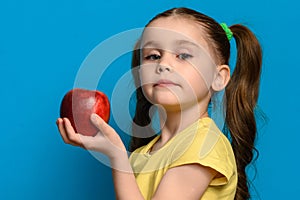 The girl stands in profile on a blue background and holds a large red apple in one hand.