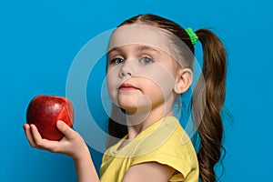 The girl stands in profile on a blue background and holds a large red apple in one hand.