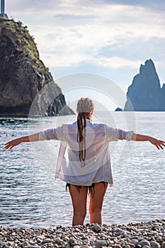 The girl stands on the shore and looks at the sea. Her hands are raised up. She wears a white shirt and her hair is in a