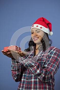 Girl staring holds a small christmas gift. she is wearing a lumberjack shirt and a christmas hat on a purple background