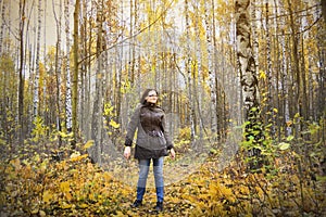 Girl stays in autumn forest among birch trees and yellow leaves.