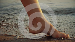 Girl stepping on wet sand of shoreline. Legs of young woman going along ocean beach during sunset. Young woman walking along the w