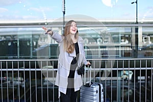 Girl stopping taxi with raised hand, valise and smartpone near airport.