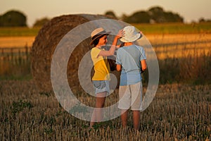 A girl in a straw hat puts a white panama on boy standing backwards, children stand in field, coil of hay behind them