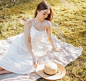 Girl with a straw hat in the spring in the park. Brunette with long hair sitting on a plaid on a background of summer nature.