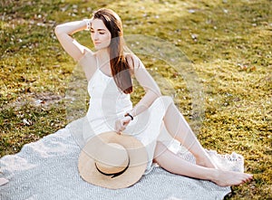 Girl with a straw hat in the spring in the park. Brunette with long hair sitting on a plaid on a background of summer nature.