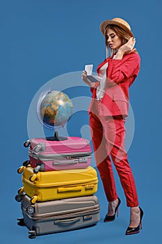 Girl in straw hat, white blouse, red pantsuit, black heels. Holding passport and ticket, posing on blue background