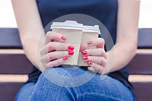 Girl on the street sits on a bench during the day. in hands with red manicure holds 2 disposable cups with tasty aromatic coffee.