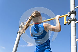 Girl on street workout. She pull-ups herself up on bar on sports ground in park. Photo from the back. Man is unrecognizable