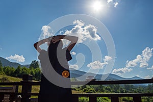 The girl stretches, looking at the morning sun and mountains. Next to her, on the railing of the balcony, is a cup of coffee