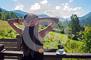 The girl stretches, looking at the morning sun and mountains. Next to her, on the railing of the balcony, is a cup of coffee