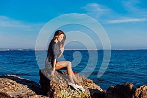 The girl in the striped dress sits on the rocks by the sea