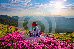 The girl in striped plaid is sitting on the lawn among rhododendrons.