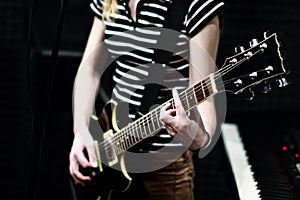 A girl in a striped shirt with an electric guitar stands on the recording Studio. The guitarist plays the strings of a musical