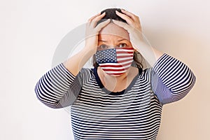 A girl in a striped sweater and medical mask painted like the flag of USA in fear covered her head with her hands.