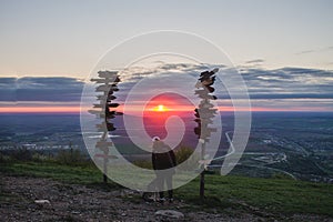 Girl stroking dog on top of mountain looking at the sunset and the plain, next to the wooden signs of the cities.