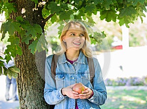 Girl student with backpack hold apple while stand near tree. Healthy snack. Students life concept. Take minute to relax