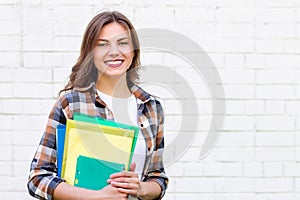 Girl student holds folders and a notebook in her hands and smiles on a background of a white brick wall