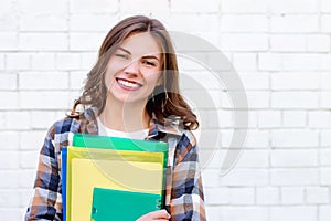 Girl student holds folders and a notebook in her hands and smiles on a background of a white brick wall