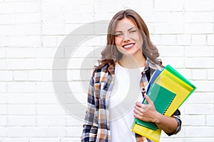 Girl student holds folders and a notebook in her hands and smiles on a background of a white brick wall