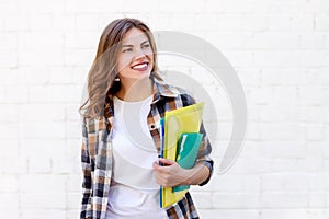 Girl student holds folders and a notebook in her hands and smiles on a background of a white brick wall