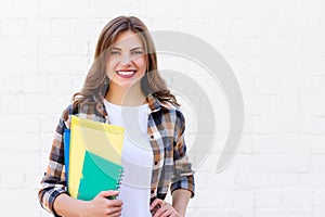 Girl student holds folders and a notebook in her hands and smiles on a background of a white brick wall