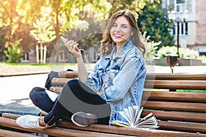 Girl student sits on a bench in the park and holds mobile phone. Girl listens to an audiobook in the park