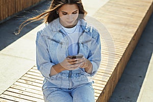 Girl student sits in public park and uses mobile phone. Beautiful brunette woman is texting with friends