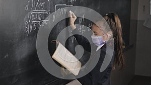 A girl student solves equations at the blackboard in a medical mask.